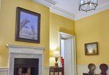 A room with a marble fireplace with a painting of a bridge hung over it. Walls are painted yellow.