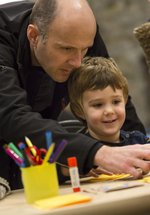 A man looks over the shoulder of a boy, he is pointing to materials on a craft table. 