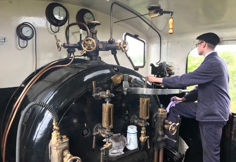 A young person wearing a cap and overalls standing in front of rods and levers on the foot plate of a steam train
