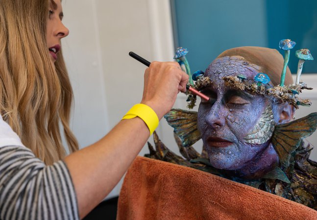A photograph of a make up artist putting special effects makeup on a model. The model's skin is blue with fins and gills. 