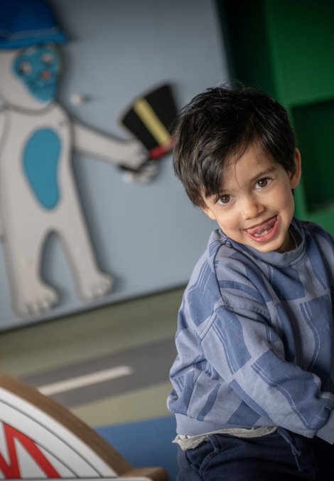 A small child looks at the camera, tongue sticking out. They are sat down, holding on to a toy, plastic steering wheel. In the background is a model of a bridge and an illustration of a cat holding a hat