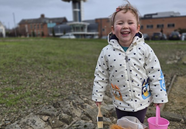 Young child smiling, on the fort site with a brush, bucket and puppet