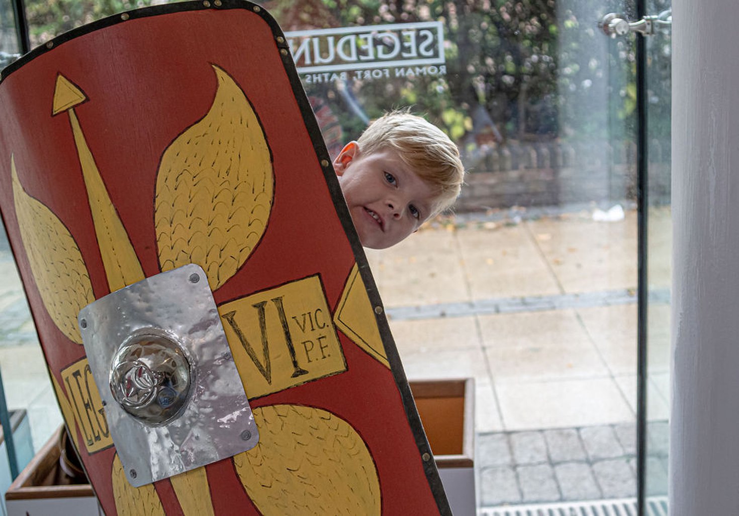 Young boy peeping out from behind a Roman style shield in the shop