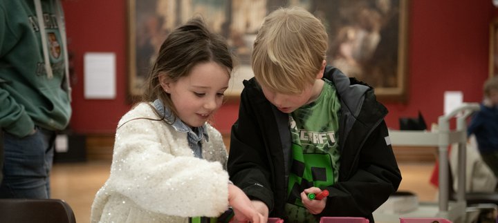 Two children looking inside a box of pens on a table, in front of a large historic painting. 