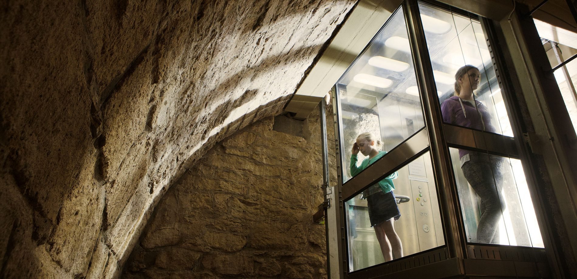 Two children in a glass lift look out at the stone wall of Hexham old Gaol's dungeon
