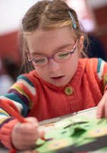 A photograph of a young girl colouring. She wears a striped jumper and glasses.