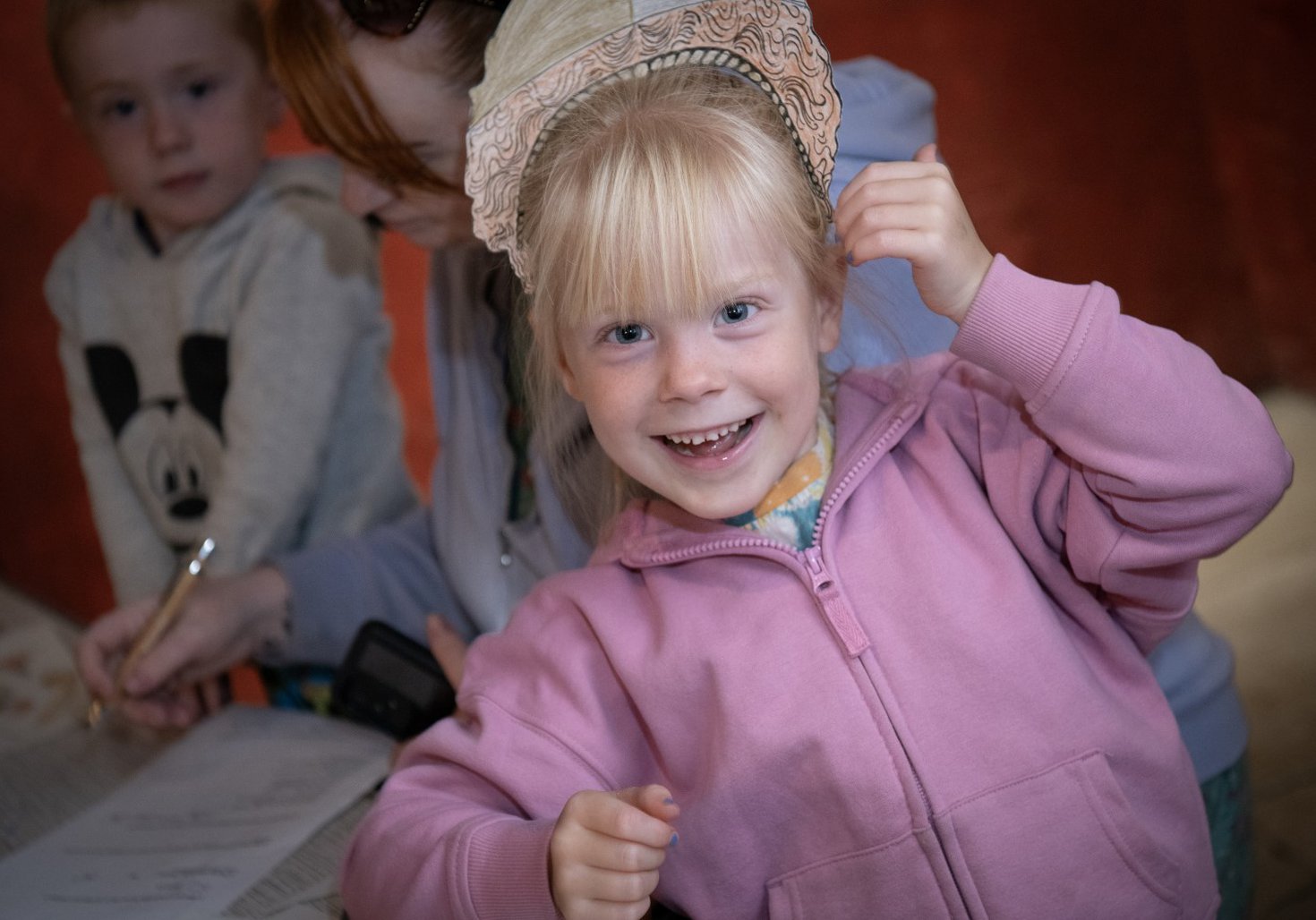 A child is wearing a paper helmet and smiling at the camera