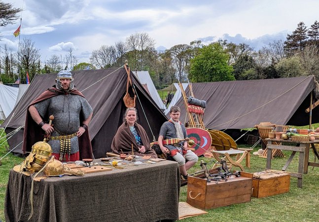 Historical reenactment camp with people in period costumes, tents, shields, helmets, and wooden tables with various items
