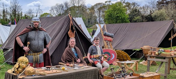 Historical reenactment camp with people in period costumes, tents, shields, helmets, and wooden tables with various items