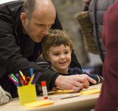 A man looks over the shoulder of a boy, he is pointing to materials on a craft table. 