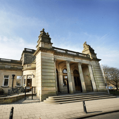 A photograph of the Shipley Art Gallery, which is made of stone and the exterior has columns on the front and two statues on the roof.