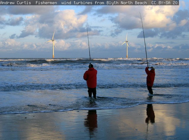 Two fishermen on the beach looking out at two offshore turbines