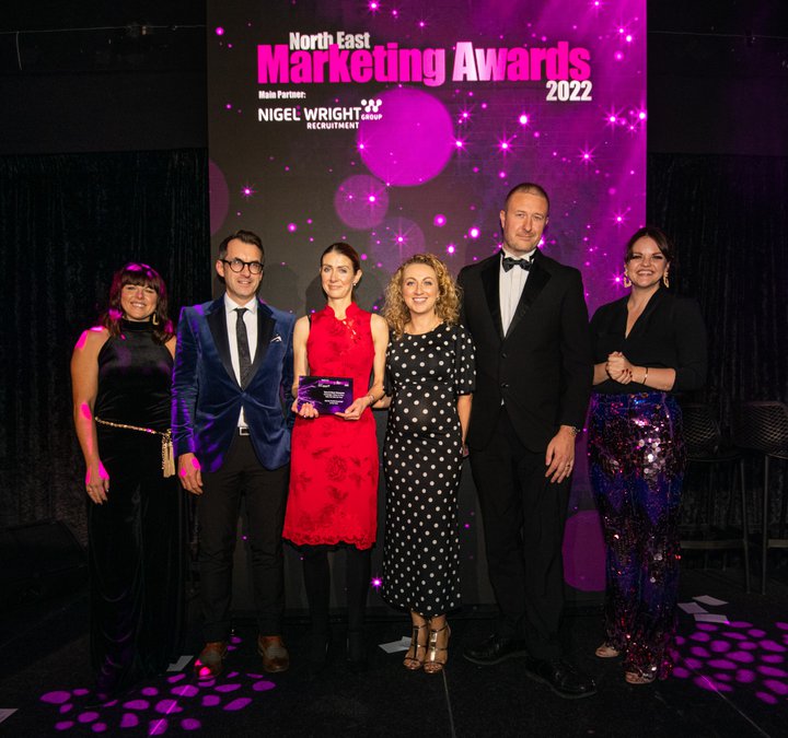Six people in evening dress receiving an award with a purple screen behind them with the words North East Marketing Awards