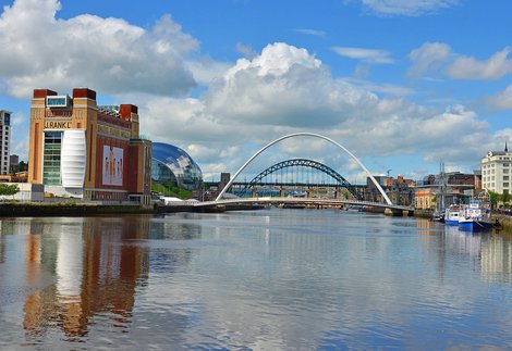A large grey bridge and a large green bridge over a sunny river. To the left is a large red and yellow brick building which reads 'Baltic Flour Mills'.
