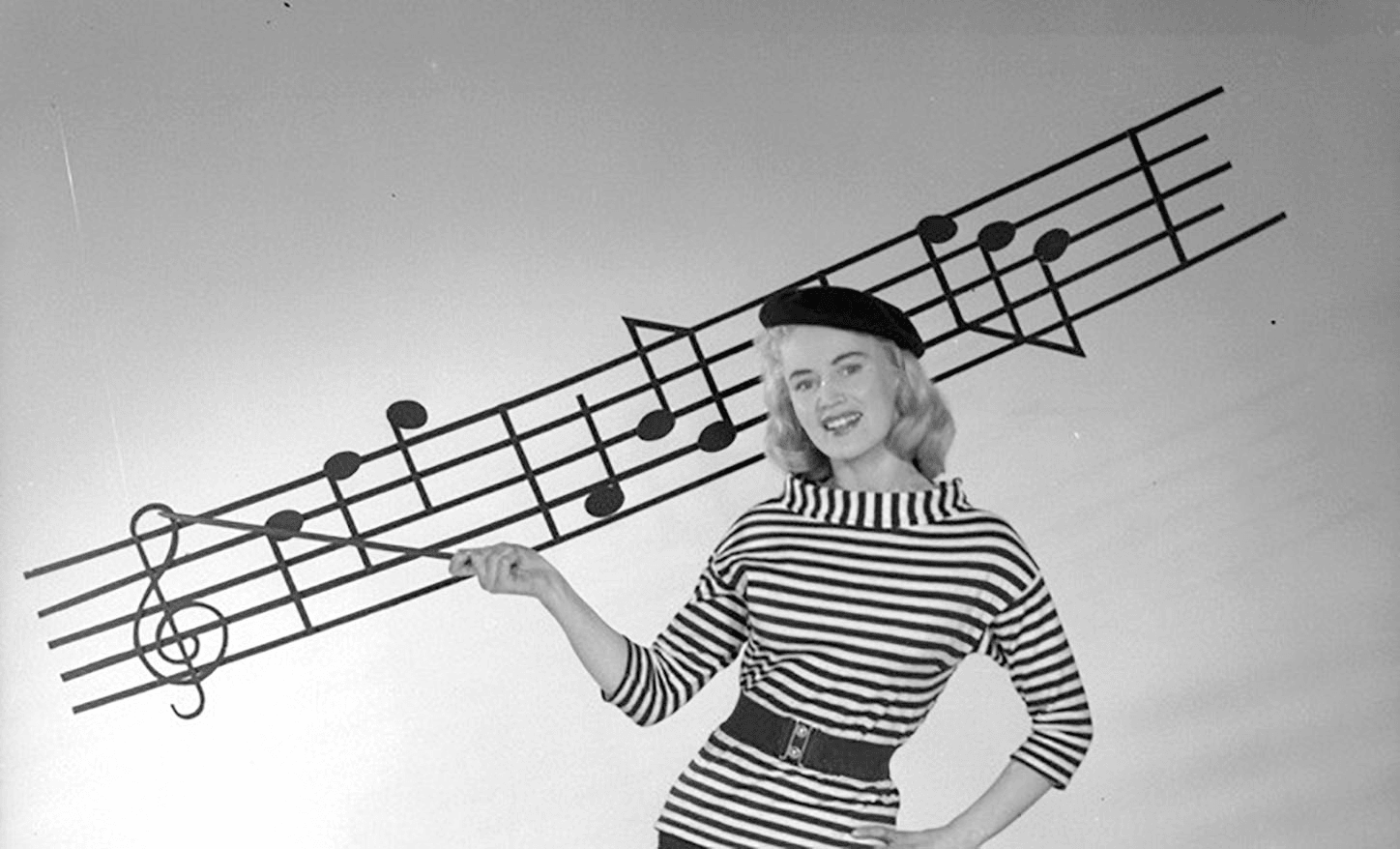 Black and white archive studio photograph. Woman in beret pointing with baton at stave of musical notation on a wall.