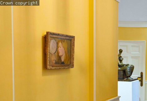 An oil painting of a woman holding a mirror. The painting is hung on a yellow wall.