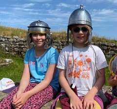Two children sitting down and wearing replica Roman soldiers helmets