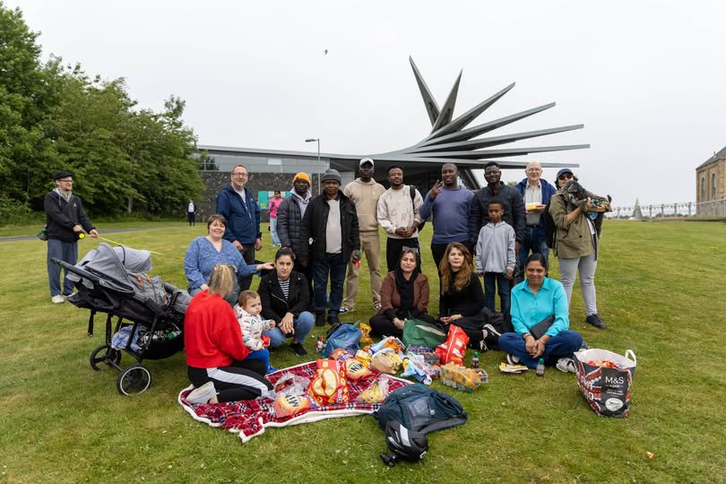 A large group of people gathered together in front of a large building with a metal angular structure of its roof. A large group of people gathered together in front of a large building with a metal angular structure of its roof.