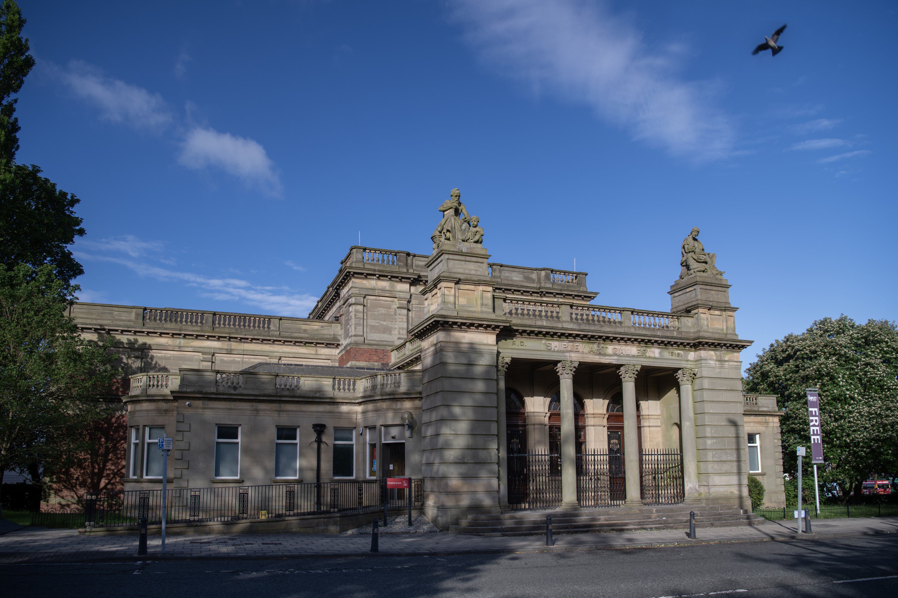 The exterior of a large stone building, with a blue sky above it. There are round columns either side of the entrance and two large pillars with statues on the top.