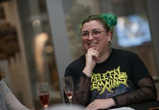 Adult person with band tee-shirt, facial piercings and green buns in hair smiles at companion