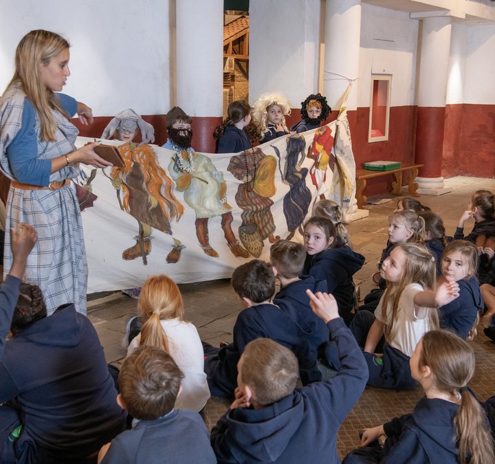 Person dressed as an ancient Brit talking to a group of schoolchildren sitting on the floor with a cloth timeline of clothing throughout the ages 