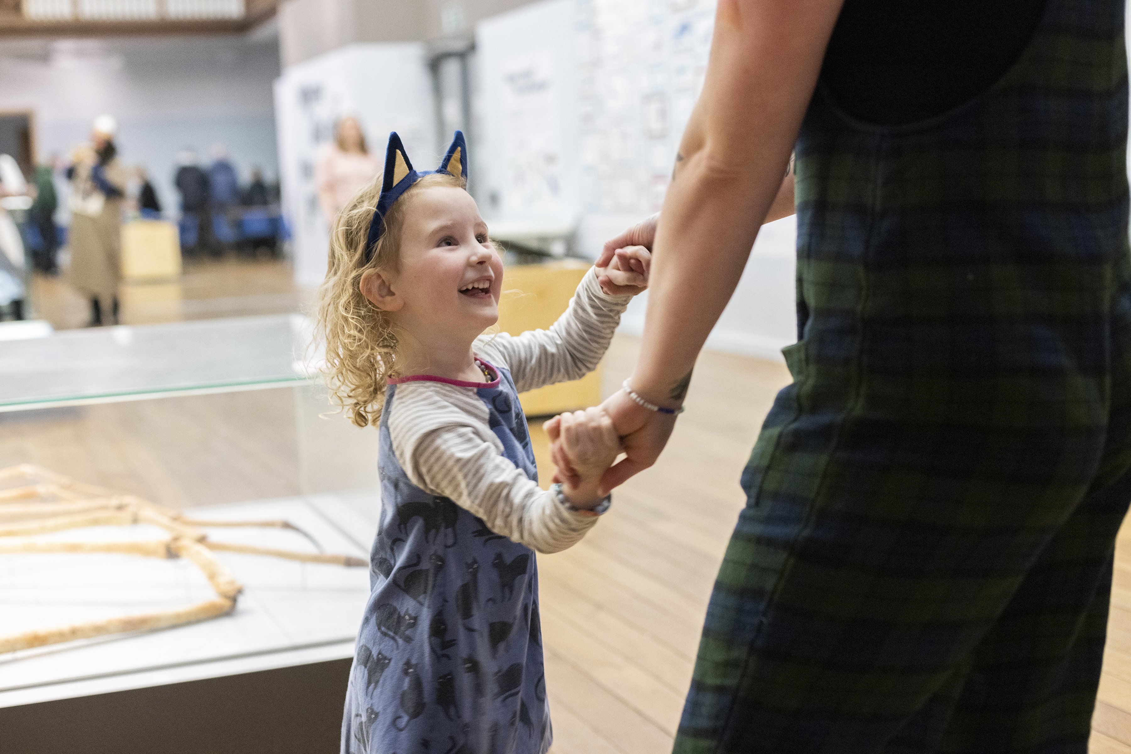 A small child wearing cat ears stands in a museum. They are holding hands with a parent and looking up at them smiling and laughing. A small child wearing cat ears stands in a museum. They are holding hands with a parent and looking up at them smiling and laughing.