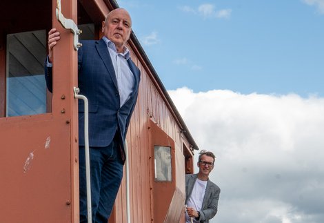 Two men standing looking out of the doorways of a red train carriage
