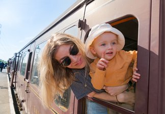 Woman with blonde hair and wearing sunglasses and her small child with mop hat leaning out of the window of a railway carriage