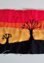 An embroidered landscape with trees silhouetted against a red, orange, and yellow sky. 