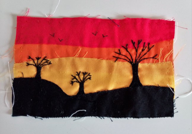 An embroidered landscape with trees silhouetted against a red, orange, and yellow sky. 