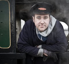 Man wearing railway cap with badge standing on the footplate of a steam engine
