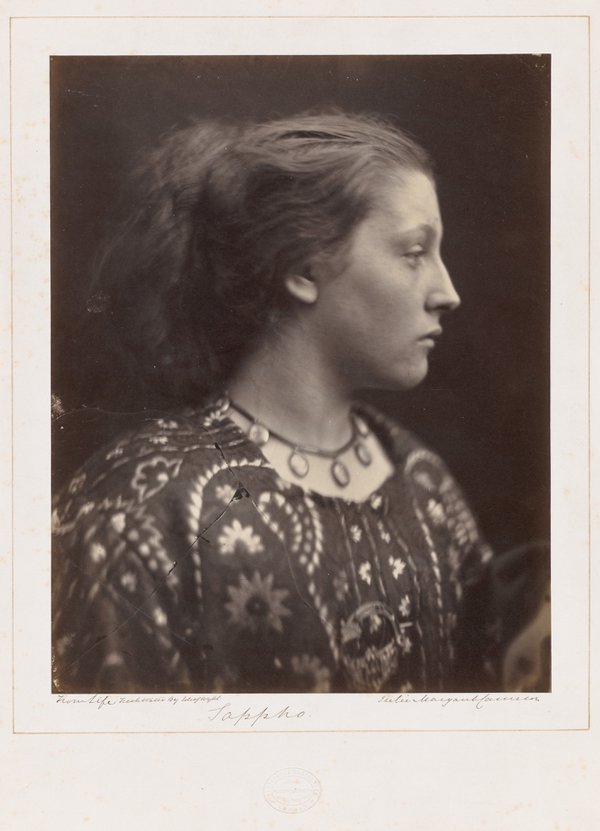 A black and white photograph of a woman’s head and shoulders in profile. She has long dark hair arranged off her face and wears a patterned dress and necklace featuring circular decorations.