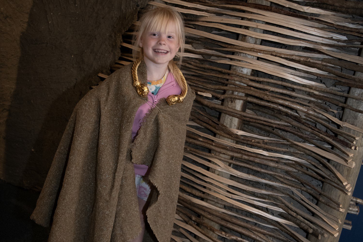 A child dressed in a Roman cloak and collar stands against a wooden backdrop and smiles at the camera A child dressed in a Roman cloak and collar stands against a wooden backdrop and smiles at the camera