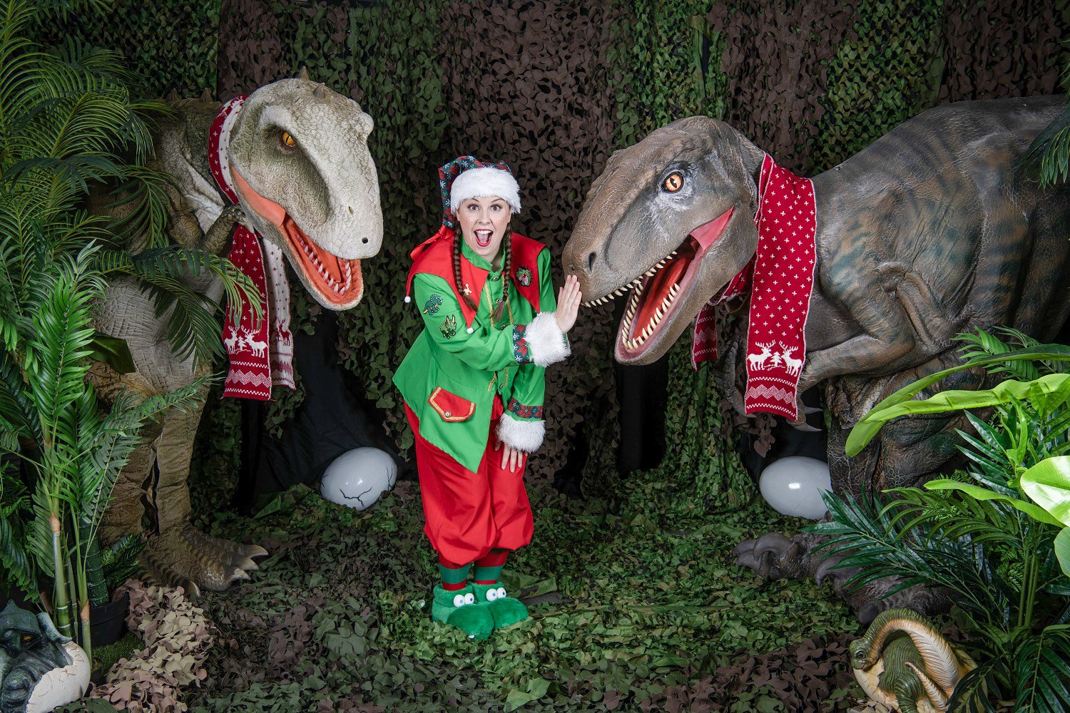 Elf standing beside two festively dressed dinosaurs.