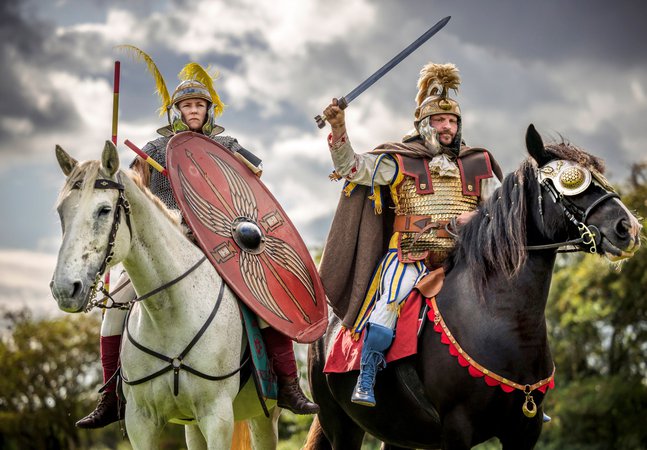 Two Roman soldiers on horseback, one with a spear and shield on a white horse, the other with a raised sword on a black horse, ready for battle under an overcast sky.