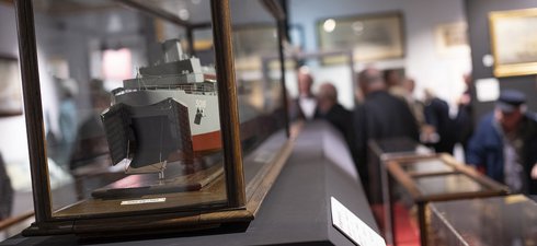 A close-up photo of a model ship. People can also be seen in the background looking at objects in the Pushing the Boat Out exhibition. 