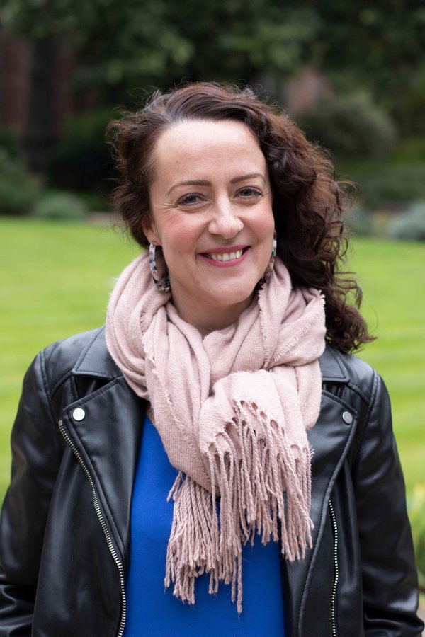 A person with dark wavy hair, smiling at the camera. They are wearing a pale pink scarf, bright blue top and black leather jacket. A person with dark wavy hair, smiling at the camera. They are wearing a pale pink scarf, bright blue top and black leather jacket.
