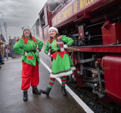 Two elves on the platform next to a steam engine with passengers in the background