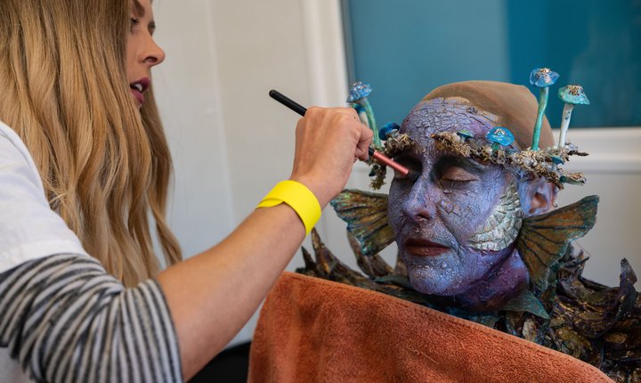 A photograph of a make up artist putting special effects makeup on a model. The model's skin is blue with fins and gills.