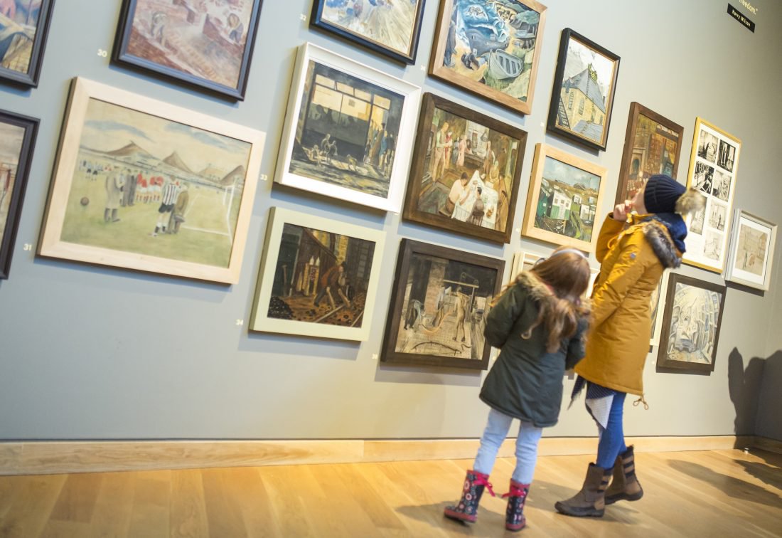 A woman and child stand in an art gallery looking at paintings displayed on the wall.