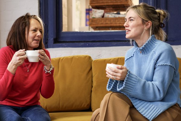 Two people smiling and drinking a hot drink together on a mustard yellow sofa.