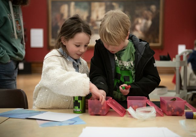 Two children looking inside a box of pens on a table, in front of a large historic painting. 