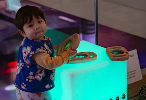 A toddler is standing next to light box cube which is placed on the ground, the toddler is holding a wooden toy.