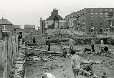 Black and white photograph of archaeologists watching the demolition of buildings while excavating a Roman fort site.