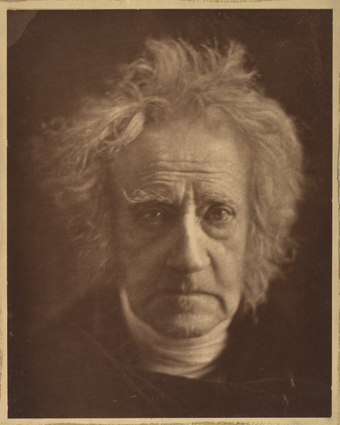 A head and shoulders sepia photograph of Sir John Herschel, a man with long white hair dressed in black with a white collar.