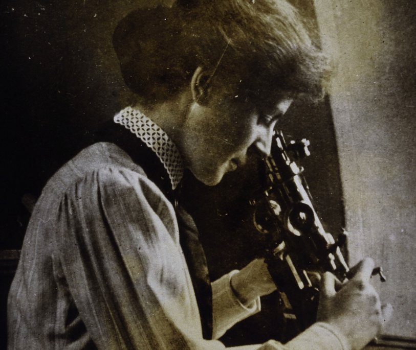 A black and white image of a woman looking through a microscope