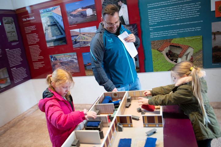 Three people, including two children and one adult, interact with a model display of the Commanding Officer's house in the reconstructed room. The background features informational panels about the fort