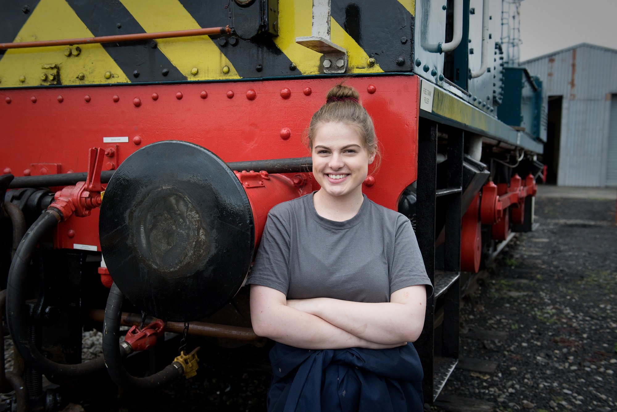 A young woman in overalls standing in front of a large diesel engine at a heritage railway.