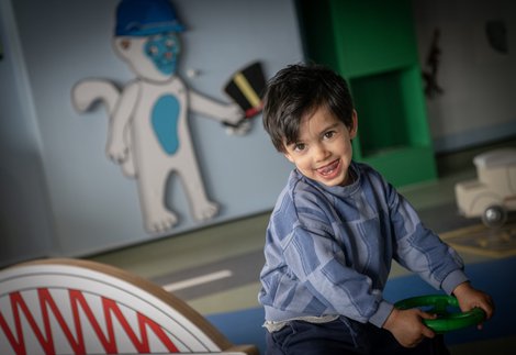 A small child looks at the camera, tongue sticking out. They are sat down, holding on to a toy, plastic steering wheel. In the background is a model of a bridge and an illustration of a cat holding a hat