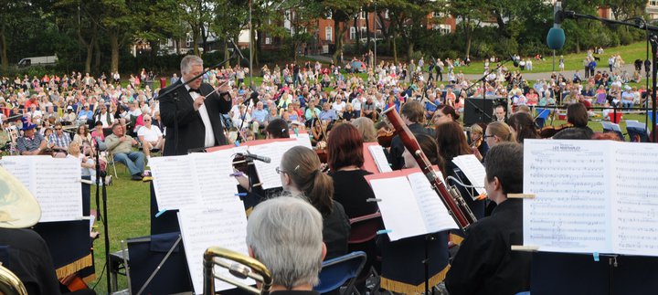 Music conductor leading an orchestra of young musicians in an outdoor performance watched by a crowd of people